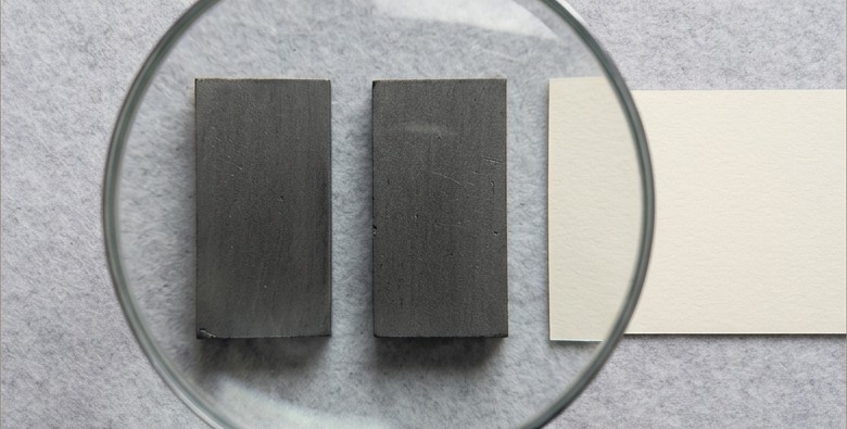 Conceptual AI illustration: Two graphite blocks viewed through an inspection lens beside blank paper.